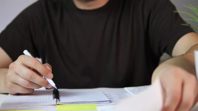 A man's hands in a casual t-shirt meticulously cross-reference a stack of receipts with entries in a notebook, carefully verifying data and recording information with a pen. This authentic scene
