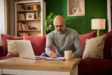 A man is seated on a red couch in a modern living room. He writes notes while reviewing financial documents on a laptop. A warm cup rests nearby, creating a calm workspace.