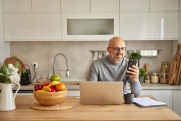 In a tidy kitchen, a man sits at a wooden table using his smartphone to review financial or insurance details. Fresh fruit fills a bowl nearby, and a laptop rests in front of him.