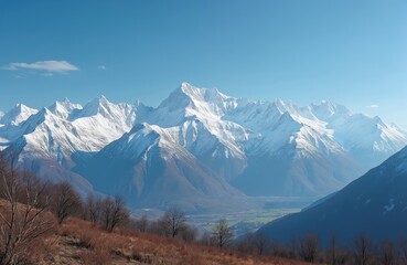 Fototapeta premium Snow covered mountain range under a clear blue sky with sparse clouds. Foreground shows dry grass and bare trees on a slope overlooking a valley.