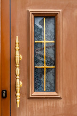 Brown front door with ornate golden door handle and rectangular window featuring gold cross pattern, classic design, close-up