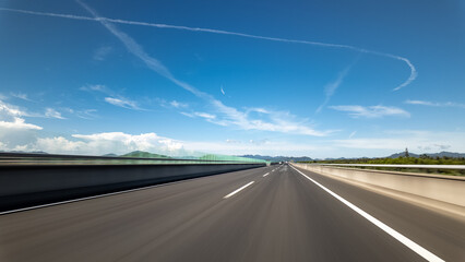 Naklejka premium Highway stretches under clear blue sky with clouds and distant mountains