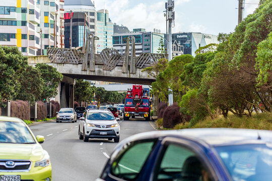 A busy Wellington street with cars passing under a uniquely designed bridge during the Crane Parade, surrounded by modern high-rise buildings and greenery