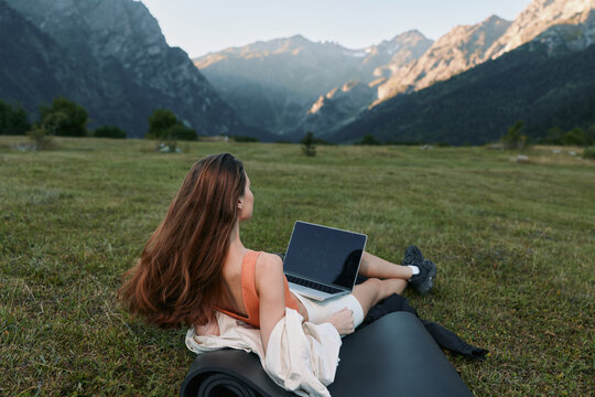 A woman in a wheelchair sits on a grassy meadow with a laptop, gazing toward distant mountains. The scene conveys focus, freedom, and a calm, productive outdoor moment.
