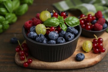 Different ripe juicy berries and basil in bowl on wooden table, closeup