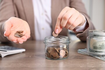 Woman putting coin into glass jar at wooden table indoors, closeup