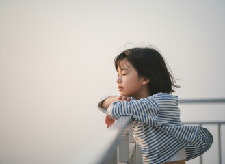 Young child peacefully leaning on balcony railing in soft morning light, eyes closed, wearing striped long sleeve top outdoors