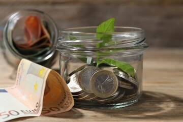Glass jar with coins, sprout and banknotes on wooden table, closeup