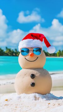 A festive sandman in a red hat and sunglasses stands on a sunny beach by calm turquoise water during Christmas holiday season