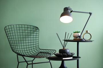 Stylish lamp, decor and reed diffuser on nesting tables near armchair against green wall indoors