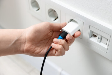 Woman plugging USB adapter with cable into electric socket on white wall, closeup