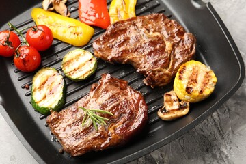 Pan with grilled beef steaks, mushrooms and vegetables on grey textured table, above view