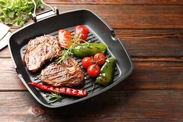 Pan with grilled beef steaks, rosemary and vegetables on wooden table, closeup. Space for text