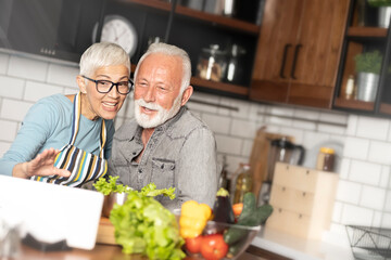 Senior couple cooking together in kitchen, using tablet to communication with their friends. Seniors, lifestyle, technology, bonding