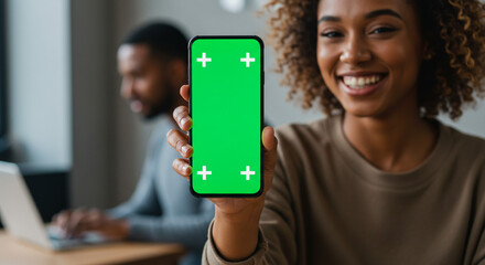 Confident woman holds green screen phone, colleague works on laptop in modern office setting