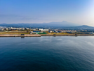 Szene am Hafen der Jeju-Insel: Schiffe, Kräne und Küstengebäude prägen das maritime Bild, eingerahmt von klarer Sicht und ruhigem Wasser. Südkorea.