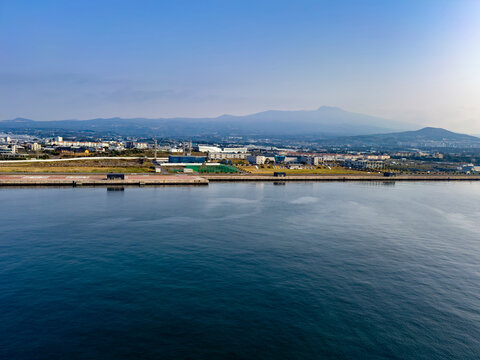 Szene am Hafen der Jeju-Insel: Schiffe, Kr&auml;ne und K&uuml;stengeb&auml;ude pr&auml;gen das maritime Bild, eingerahmt von klarer Sicht und ruhigem Wasser. S&uuml;dkorea.
