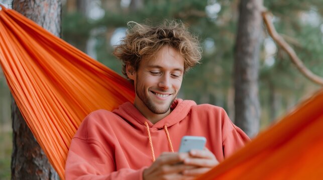 Young man relaxing in an orange hammock while using smartphone in a serene forest setting during the day - Powered by Adobe