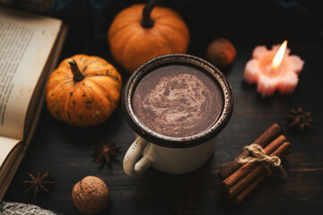Cozy autumn scene with mug of hot chocolate, book, pumpkins, spices and candles on rustic dark wooden background, warm seasonal atmosphere