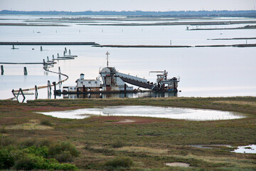 Industrial dredging machinery operates in a quiet coastal lagoon or marshland.
