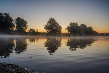Stimmungsvolle Morgenaufnahme am Fluss mit nebligem Wasser und Bäumen im Licht der Morgendämmerung
