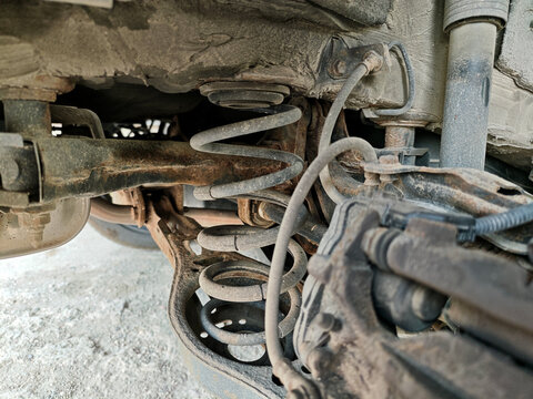 Close-up of a dirty and rusty car undercarriage showing the coil spring suspension, shock absorber, and brake caliper during a mechanical inspection.
