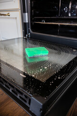 Oven door is lowered down and washed. Sponge for washing lies on its surface