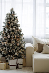 A large Christmas tree with many ornaments sits in front of a window. The room is decorated with a white couch and a few boxes. Scene is festive and cozy, with the tree and decorations creating a warm