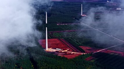 Time-lapse aerial of wind turbine towers rising among dense forests and red soil fields in Latvia. Latvia - Powered by Adobe