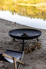 Relaxing by the River: A Cozy Fire Pit, Logs, and Camping Chair Ready for a Perfect Evening Outdoors by the Water, Surrounded by Nature's Calm Serenity