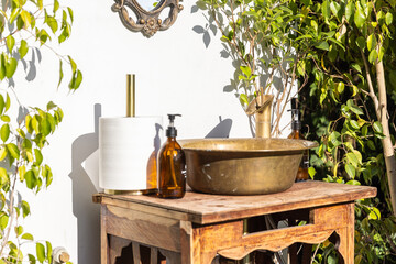 Outdoor Rustic Bathroom Setup Featuring a Vintage Brass Sink with Wooden Base Surrounded by Lush Greenery and Decorative Mirror on a Sunlit Wall