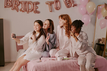 Group of four young adult women sitting on bed taking selfie with smartphone, smiling and making peace signs during bachelorette party celebration