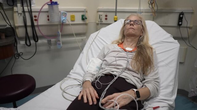 A mature woman lies in a hospital bed while connected to heart and EKG monitors in the emergency room. She appears to be in a state of rest and monitoring.