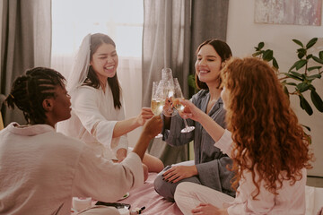 Group of four young adult women, multiethnic, sitting together and smiling while clinking champagne glasses during bachelorette party celebration in bright indoor setting