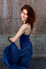 Fototapeta premium Smiling Woman with Red Hair Twirls Blue Skirt Against Granite Wall