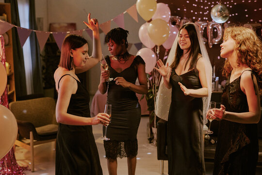 Group of young adult women dancing and holding champagne glasses at bachelorette party, one woman wearing bridal veil, balloons and decorations visible - Powered by Adobe