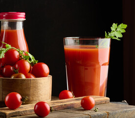 Red cherry tomatoes in a wooden bowl. Tomatoes are scattered on a wooden board, two bottles of tomato juice and a glass are standing nearby.