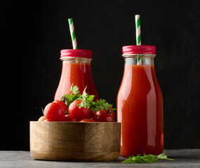 Glass bottles with tomato juice on black table and wooden bowl with cherry tomatoes