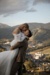 Fototapeta premium A beautiful, happy young couple in love on a wedding walk and photo shoot high in the mountains. The groom in a jacket and suit and the bride in a white dress at a resort. Kissing and hugging