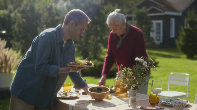 Zoom out shot of middle-aged Caucasian man bringing fruit platter and salad bowl, helping his senior mother to set beautiful festive table for family gathering in cozy backyard on nice summer day