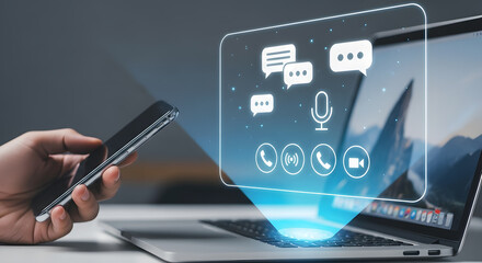 A hand holding a smartphone projecting a holographic interface with communication icons, including chat bubbles, a microphone, and video call symbols, towards a laptop screen.