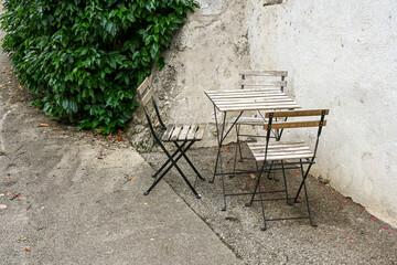 A simple set of weathered wooden bistro chairs and a table placed against a textured wall in a quiet corner of France, evoking a rustic and peaceful atmosphere.