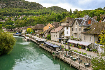 A charming riverside view of Chanaz, France, with traditional houses, waterfront caf&eacute;s, and lush green hills in the background.