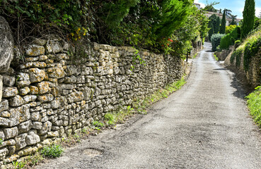 A rustic stone wall lining a quiet countryside lane in Bonnieux, France, surrounded by greenery and traditional Provençal charm