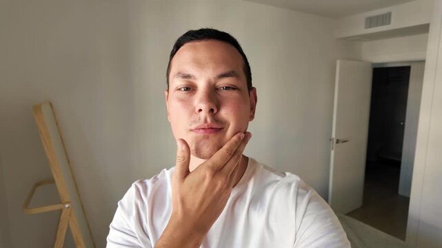 A man with noticeable hair loss is seated on a bed in a light-colored room. His casual white t-shirt contrasts with the neutral background. The scene captures an intimate moment of reflection.