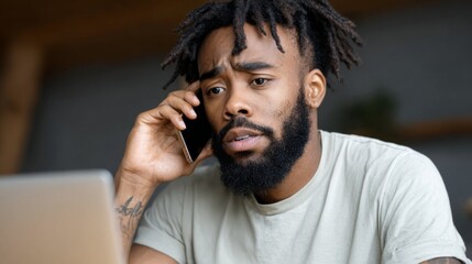 Man engaged in a serious phone conversation while working on a laptop at home during the day