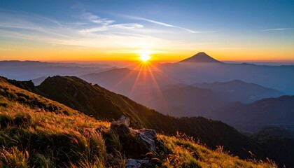 Obraz premium Sunrise over mountain range with radiating sun rays. Hillside with grass and rocks in the foreground. Distant, shadowed mountains