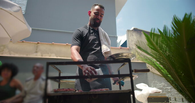 Man smiling while grilling barbecue outdoors, holding seasoning jar and knife, casual backyard setting with family gathering in background, summer social event