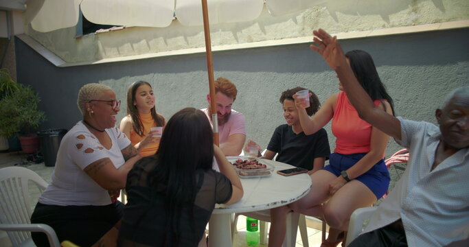 Group of diverse friends and family raising glasses in toast at outdoor barbecue, celebrating unity and joy around a table under a large umbrella in a vibrant backyard