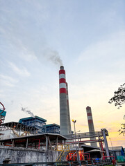 Industrial plant featuring tall chimney emitting smoke against a sunset colored sky landscape view.
